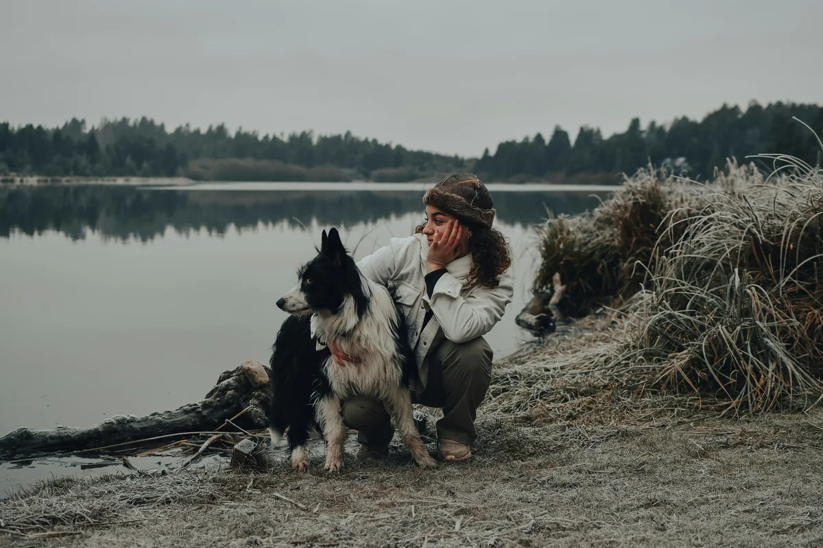 man in gray jacket and black pants sitting beside black and white dog on brown field - human names for dogs